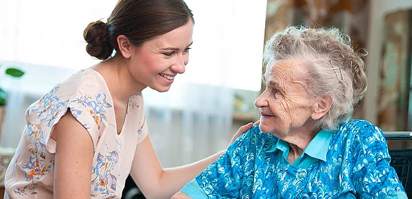 Foto einer jungen Frau, die lächelnd einer Seniorin die Hand auf die Schulter legt
