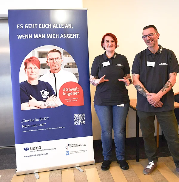 Pflegefachkräfte und Deeskalationstrainer Britta Graf und Jörg Hinderlich vor einem Plakat mit ihren Porträts.