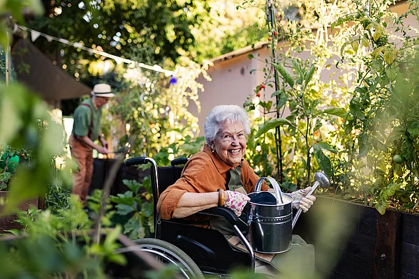 Eine ältere Dame im Rollstuhl gießt ein Hochbeet, im Hintergrund arbeitet ein Mann.