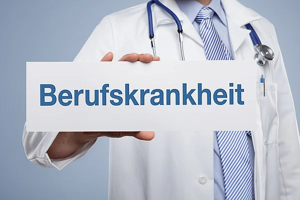 Ein Mann im Arztkittel halt ein Schild mit der Aufschrift “Berufskrankheit” vor sich.
