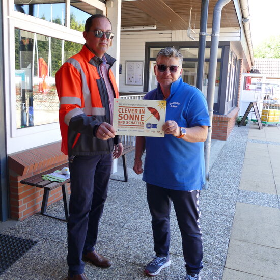 Frau in blauem T-Shirt und Mann in Arbeitsschutzkleidung zeigen Schild mit Auszeichnung "Clever in Sonne und Schatten"