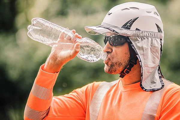 Ein Bauarbeiter mit Schutzhelm, Sonnenbrille und Nackentuch trinkt Wasser in der Pause.