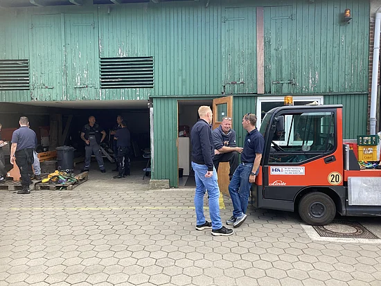 Drei Männer in orangeroter Arbeitskleidung im Gespräch vor einer Werkshalle