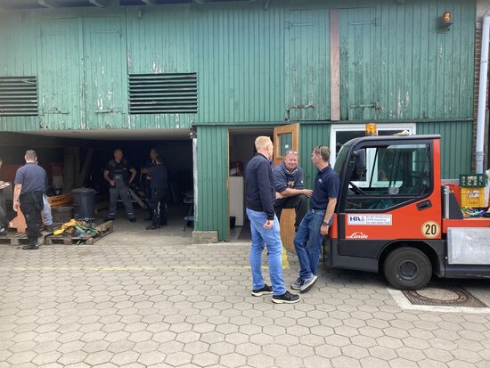 Drei Männer in orangeroter Arbeitskleidung im Gespräch vor einer Werkshalle