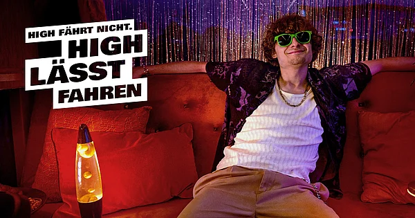 Ein junger Mann sitzt entspannt auf einem roten Sofa in einem Raum mit Retro-Atmosphäre. Er trägt ein weißes Unterhemd, eine offene, gemusterte Hemdjacke, beige Hose, eine goldene Kette und auffällige grüne Sonnenbrillen. Die Beleuchtung ist warm und stimmungsvoll. Neben ihm steht eine gelbe Lavalampe auf einem Tisch. Der Hintergrund ist mit schimmernden, lila-blauen Vorhängen dekoriert. Überlagert ist ein großer weißer Schriftzug: „High fährt nicht. High lässt fahren.“