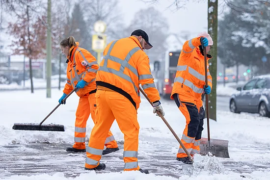 Drei Beschäftigte der Stadtreinigung Hamburg in orangefarbener Sicherheitskleidung schieben Schnee.