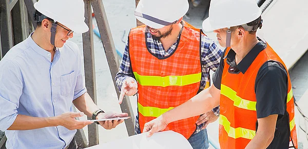 Drei Männer mit Warnwesten und Bauhelmen besprechen sich auf einer Baustelle.