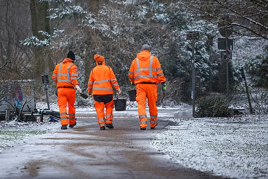 Drei Beschäftigte der Stadtreinigung Hamburg in orangefarbener Sicherheitskleidung streuen im Winter abstumpfendes Material auf einen Gehweg.