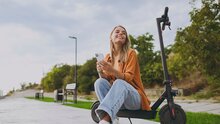 Eine junge Frau mit Smartphone pausiert sitzend auf ihrem E-Roller.