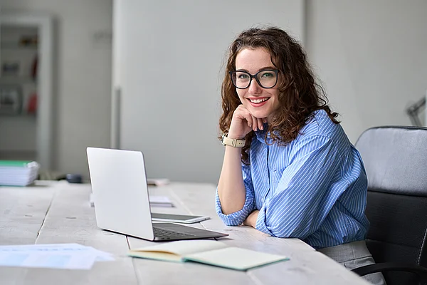 Eine junge Frau mit Brille sitzt lächelnd vor einem Laptop.