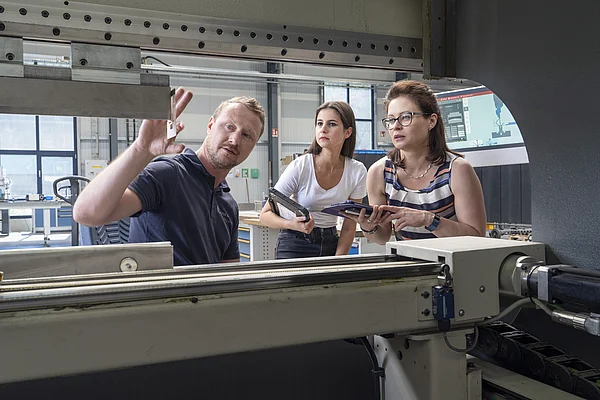 Ein Mann erklärt in einer Werkstatt zwei Frauen eine Maschine.