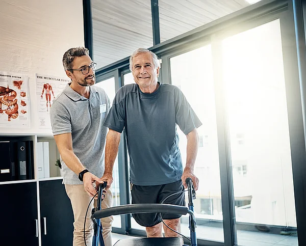 Physiotherapeut stützt älteren Mann beim Gehen am Rollator.