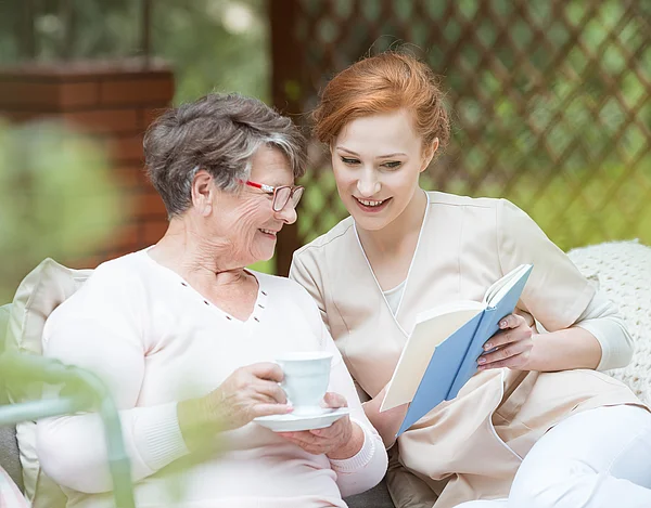 Eine jüngere Frau liest einer älteren aus einem Buch vor.