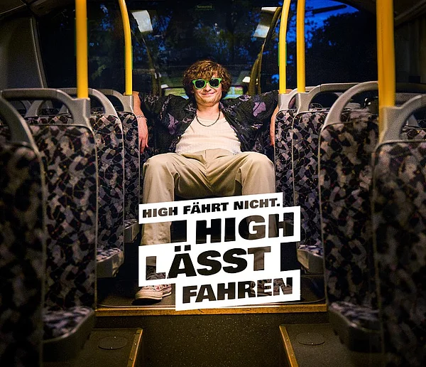 Ein junger Mann sitzt entspannt auf einem roten Sofa in einem Raum mit Retro-Atmosphäre. Er trägt ein weißes Unterhemd, eine offene, gemusterte Hemdjacke, beige Hose, eine goldene Kette und auffällige grüne Sonnenbrillen. Die Beleuchtung ist warm und stimmungsvoll. Neben ihm steht eine gelbe Lavalampe auf einem Tisch. Der Hintergrund ist mit schimmernden, lila-blauen Vorhängen dekoriert. Überlagert ist ein großer weißer Schriftzug: „High fährt nicht. High lässt fahren.“.