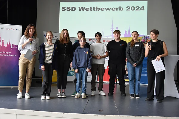 Sieben Schülerinnen bzw. Schüler und zwei erwachsene Frauen stehen auf einer Bühne.